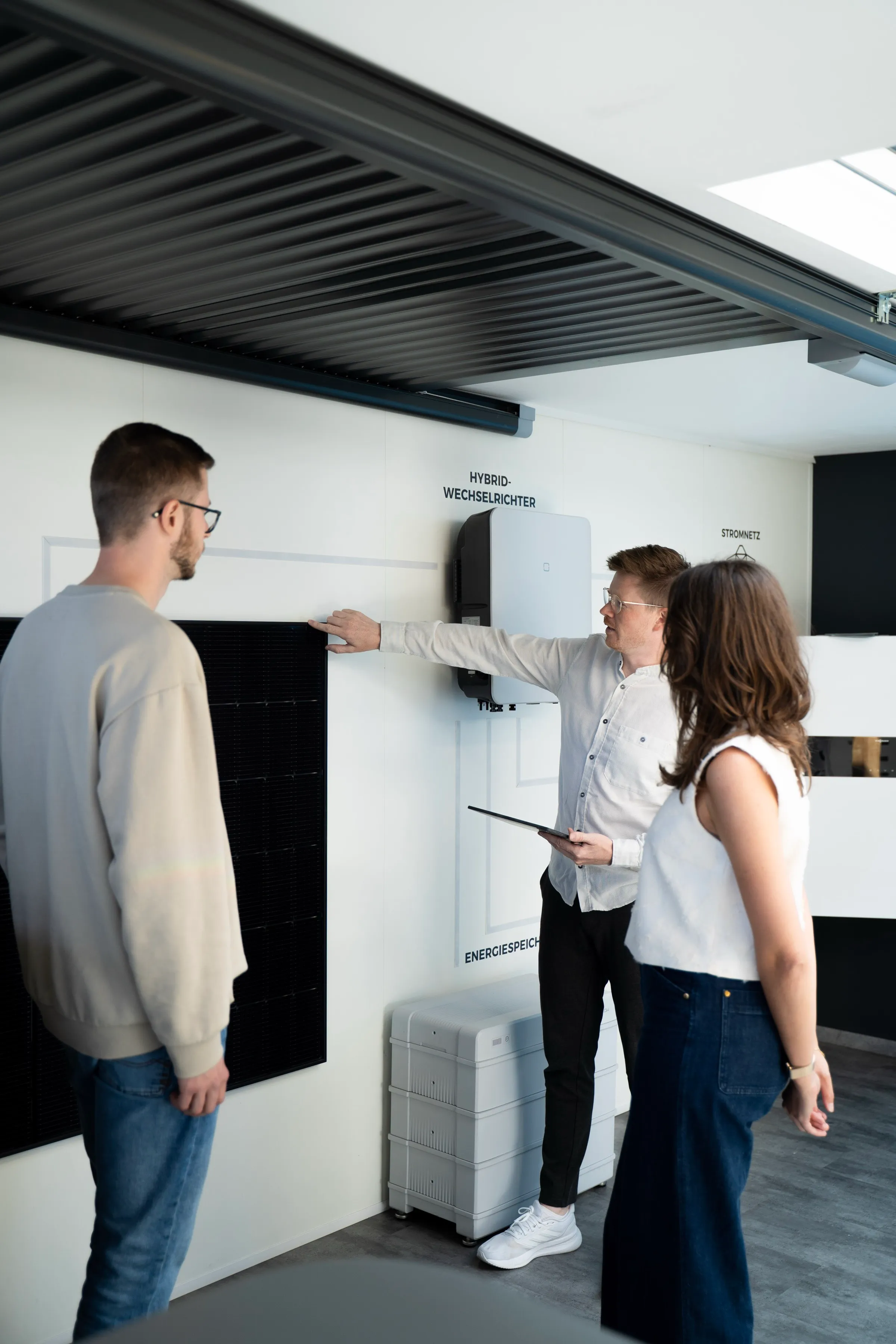 Ein Energieberater erklärt an einer Technikwand den Hybrid-Wechselrichter und den Energiespeicher; zwei Personen hören zu.